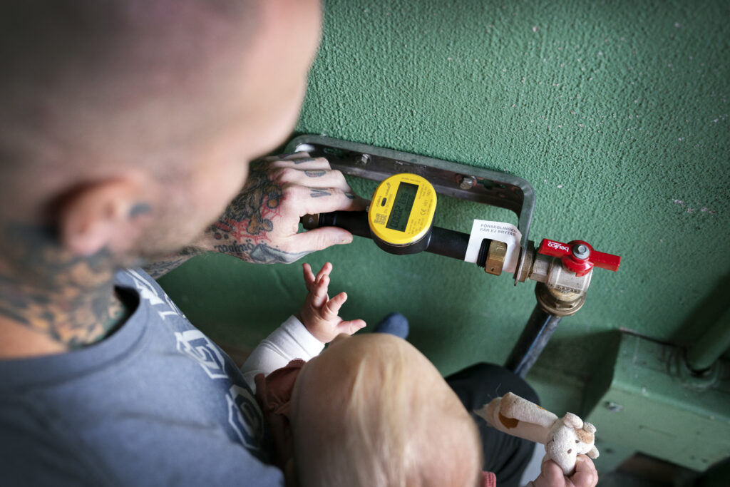 I ett garage, på en grönputsad vägg sitter en digital vattenmätare. Den är ansluten med en röd kran till en kraftig slang för inkommande vatten. Kranen är öppen. En man med ett barn i famnen har blicken vänd mot rörinstallationen. Han håller sin tatuerade hand intill vattenmätaren. Barnet sträcker sin hand mot hans.
